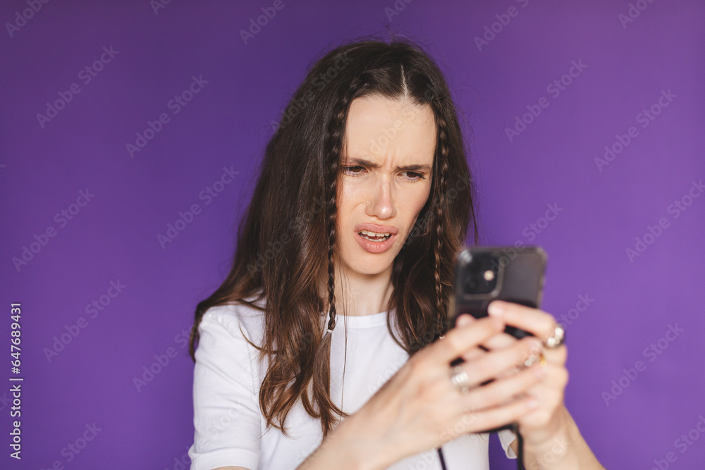 Mad and shock young brunette woman using mobile phone isolated on purple background. Yelling unhappy woman hold mobile phone. Disappointed sad upset lady horrified impressed news. WTF. Oh no. Mad and shock young brunette woman using mobile phone isolated on purple background. Yelling unhappy woman hold mobile phone. Disappointed sad upset lady horrified impressed news. WTF. Oh no.