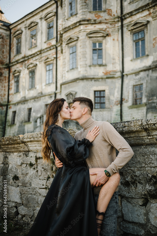 Woman in black long dress hugs man stand near ancient palace at sunset. Female kissing male in street. Luxury couple newlyweds near old Pidhirtsi Castle, Lviv Ukraine. Stylish bride and groom outdoors