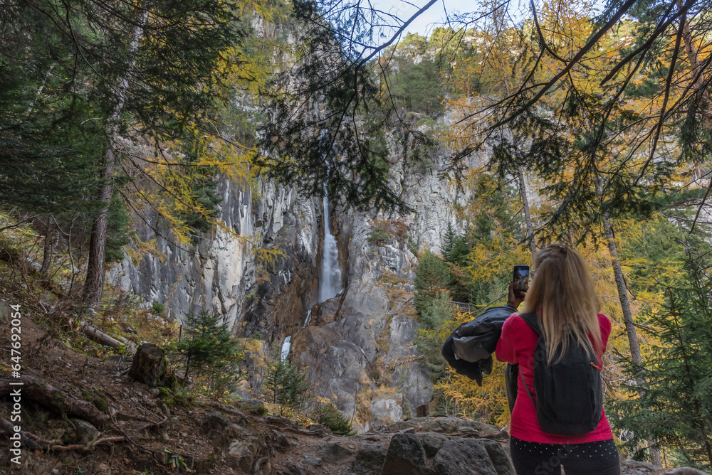 Woman with mobile phone taking photos of the waterfall on a rocky mountain in a forest.