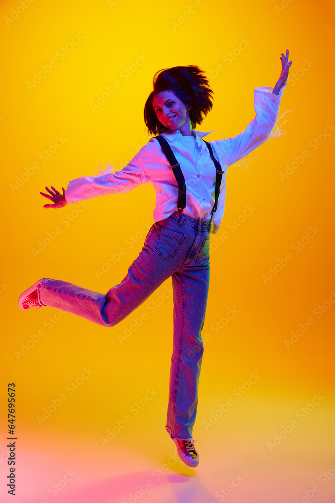 Full-length image of young woman in her 30s, wearing casual clothes and jumping against orange studio background in neon light. Concept of human emotions, fashion, lifestyle, facial expression, ad Full-length image of young woman in her 30s, wearing casual clothes and jumping against orange studio background in neon light. Concept of human emotions, fashion, lifestyle, facial expression, ad