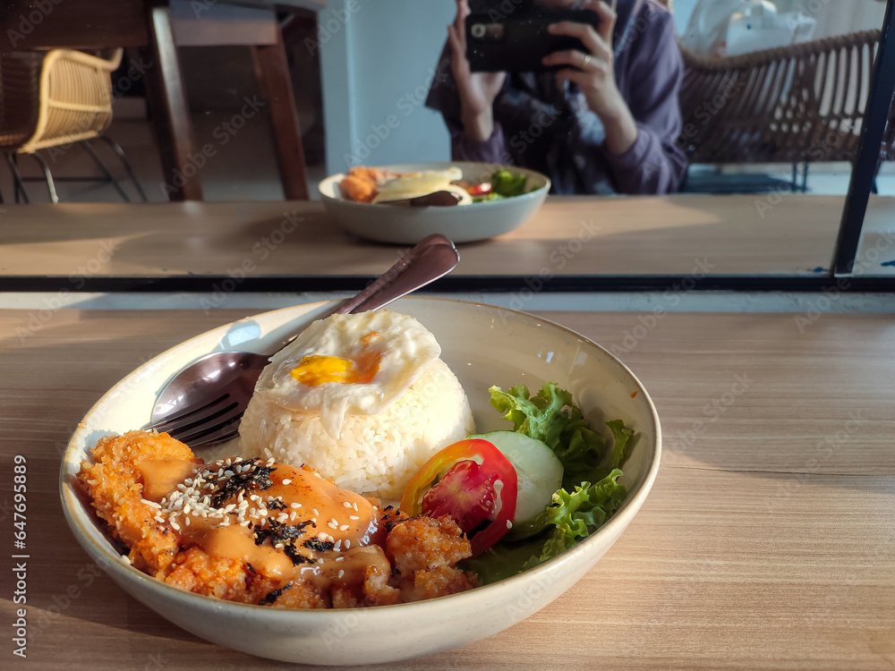 A plate of lunch with mentai chicken as a side dish with a blurry background of the reflection in the mirror of a woman who is taking photos of the food with her cellphone.