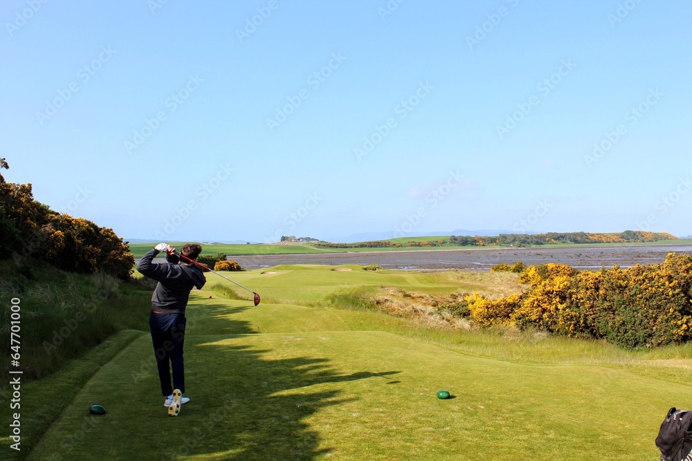 A young man swinging a golf club on a tee box surrounded by the beautiful views of the sea in the background, outside Inverness, Scotland