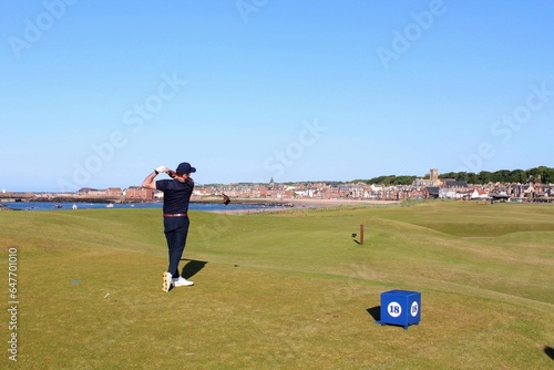 A young man swinging a golf club on a tee box surrounded by the beautiful views of the sea and North Berwick in the background, in Scotland