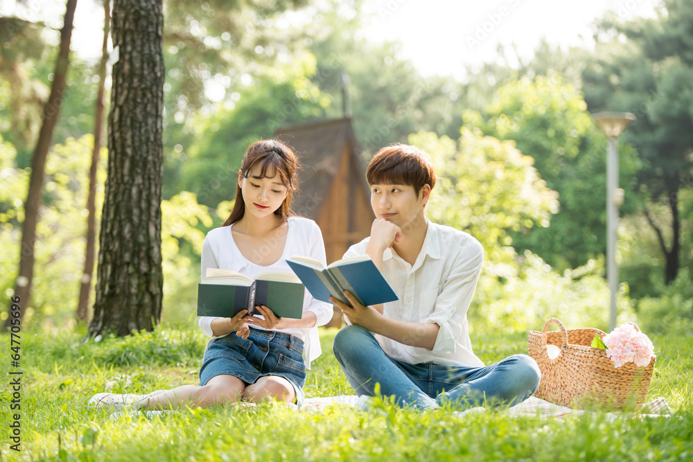 A young male and female couple are enjoying a pleasant picnic while drinking drinks and reading books on the forested park lawn. A young male and female couple are enjoying a pleasant picnic while drinking drinks and reading books on the forested park lawn.