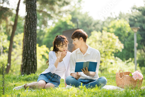 A young male and female couple are enjoying a pleasant picnic while drinking drinks and reading books on the forested park lawn.