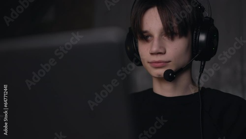 Front view of young computer gamer wearing headphones playing pc games, communicating in live stream in a dark room. Generation z, teen boy using laptop for esports or virtual sports
