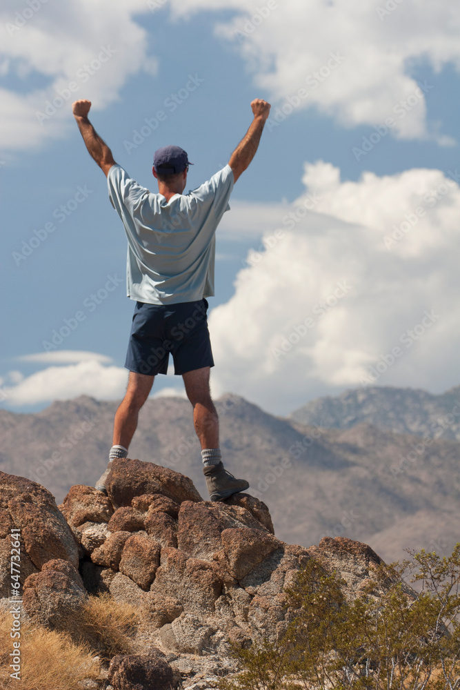 Male Hiker With Arms Raised On Top Of A Desert Mountain With Mountain Range In Distance; Palm Springs, California, United States of America Male Hiker With Arms Raised On Top Of A Desert Mountain With Mountain Range In Distance; Palm Springs, California, United States of America