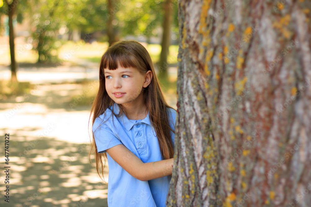 Young beautiful smiling girl nine years old in a blue t-shirt peeking out from the trunk of a big tree playing hide and seek Young beautiful smiling girl nine years old in a blue t-shirt peeking out from the trunk of a big tree playing hide and seek