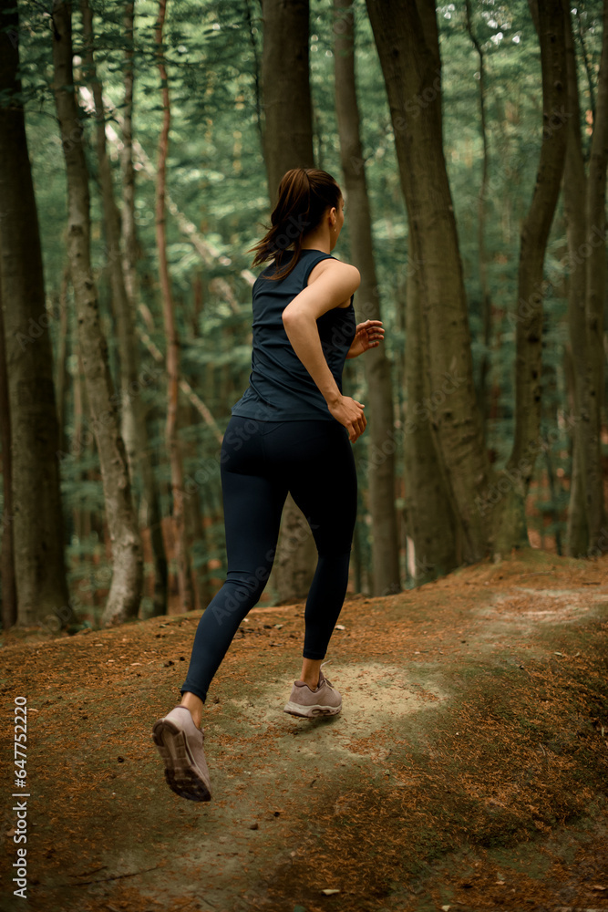 Female with brown hair in pony tail running and jogging on hills in forest between tall deciduous trees Female with brown hair in pony tail running and jogging on hills in forest between tall deciduous trees