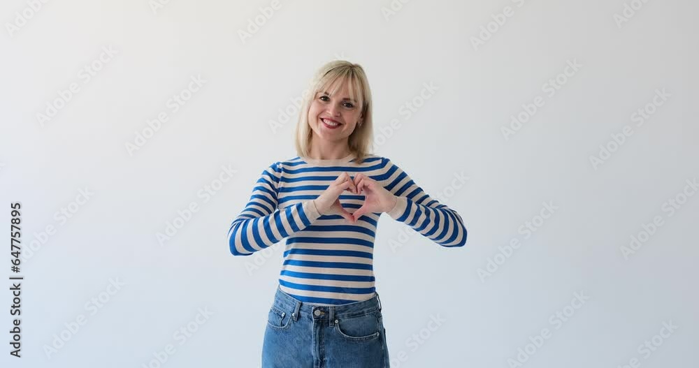 A joyful Caucasian woman is seen expressing happiness by forming a heart shape with her hands. Her beaming smile and the heart gesture symbolize love and affection. A joyful Caucasian woman is seen expressing happiness by forming a heart shape with her hands. Her beaming smile and the heart gesture symbolize love and affection.