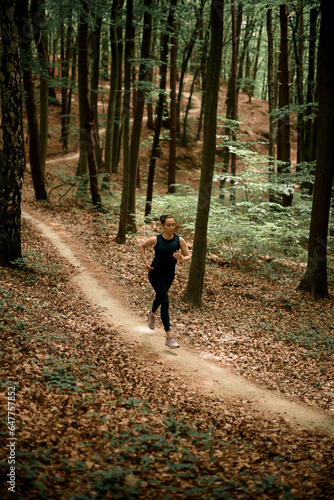 Front view of young slim woman doing daily jog on forest path between tall leafy trees