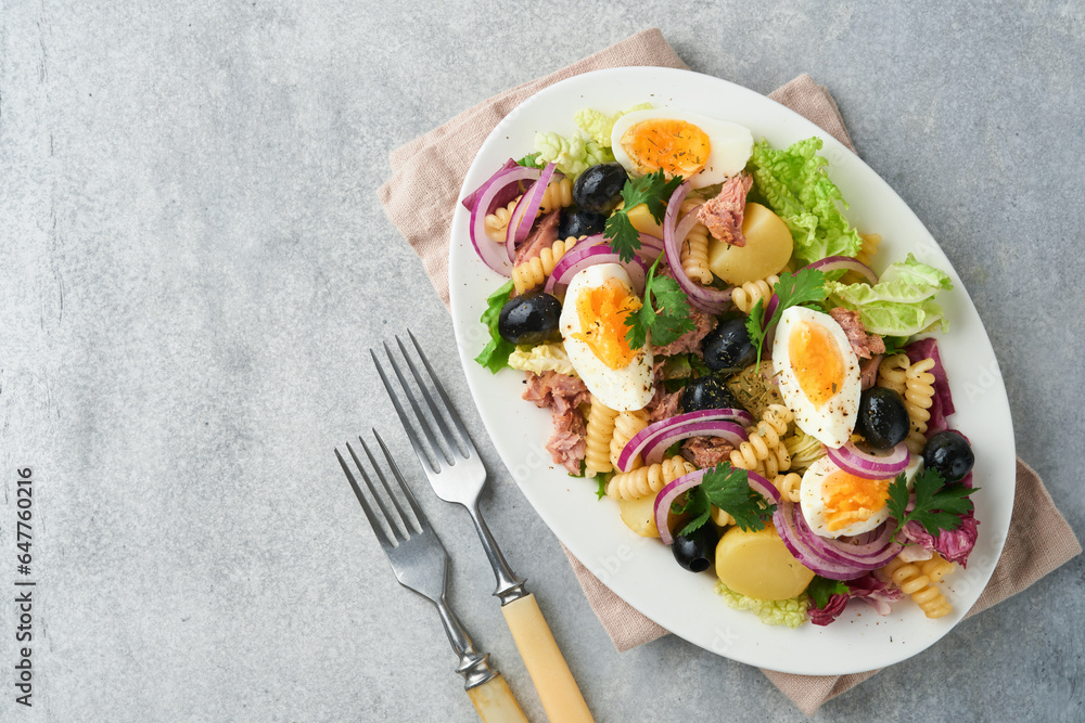 Tuna salad with pasta, eggs, potatoes, olives, red onions and sauce in white plate on old light gray concrete table background. Nicoise salad. French cuisine. Top view. Flat lay. Tuna salad with pasta, eggs, potatoes, olives, red onions and sauce in white plate on old light gray concrete table background. Nicoise salad. French cuisine. Top view. Flat lay.