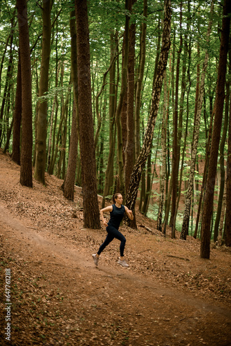 Athlete young woman in black sportswear jogging downhill on forest trail, tall leafy trees on background