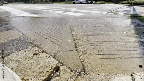 A water main break has water flowing down a handicap ramp and into the street as cars drive by in the background.