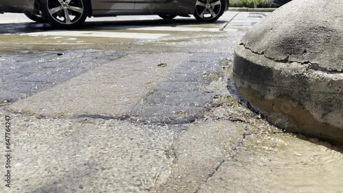 Water main leak has water flowing out from under a light pole base and into the street as cars drive by in the background.
