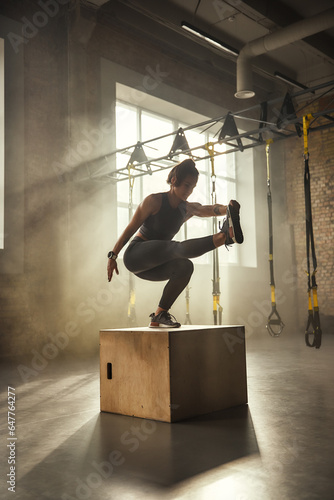 Becoming the best. Side view of athletic woman in sportswear doing squat and training legs while standing on wooden box at gym.