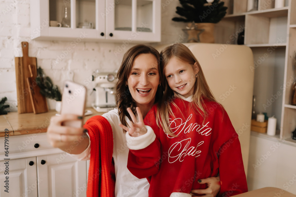 Happy mom and teen daughter in Christmas sweaters hugging, looking into a mobile phone camera, taking selfies using a mobile phone and technology on a winter holiday. Selective focus Happy mom and teen daughter in Christmas sweaters hugging, looking into a mobile phone camera, taking selfies using a mobile phone and technology on a winter holiday. Selective focus