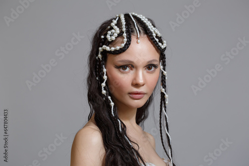 Beauty portrait of a brunette girl with a braid hairstyle with pearls on long hair isolated on a gray background.