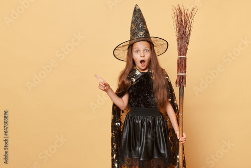 Shocked surprised girl dressed in witch costume holding broom isolated over beige background halloween celebration pointing away at advertisement area, copy space for promotion.