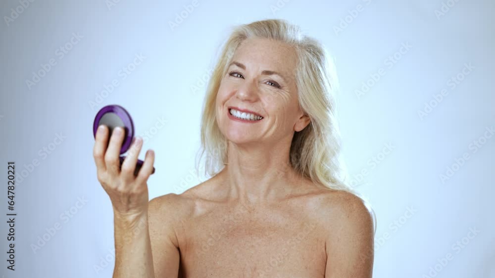 Portrait of 50s, 60s, mature middle aged woman looking at herself in compact mirror and smiling looking at the camera isolated on white background with copy space.