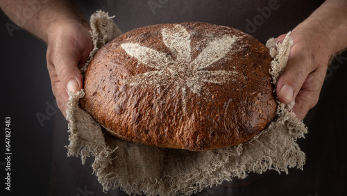 Man baker holding fresh spelt wheat hemp flour loaf of bread on rustic linen towel on dark background. Loaf bread with crushed hemp seeds decorated cannabis leaf from flour.