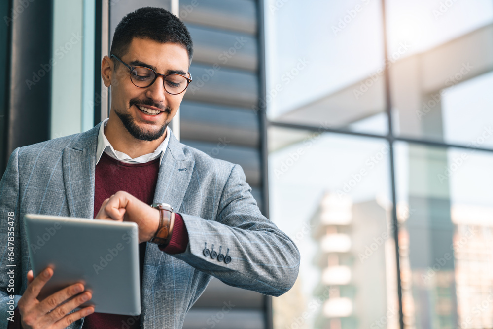 Young happy businessman looking at watch while holding digital tablet outside of modern building on a sunny day. Male CEO checking time for his next appointment. Young happy businessman looking at watch while holding digital tablet outside of modern building on a sunny day. Male CEO checking time for his next appointment.
