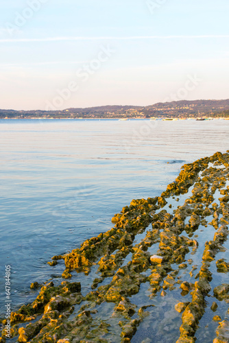 Morning rest on Lake Garda in Italy. Due to the low water level rocks and stones stick out. In the background the red, blue and purple sky, calm water, boats, buoys and the shore in the morning light