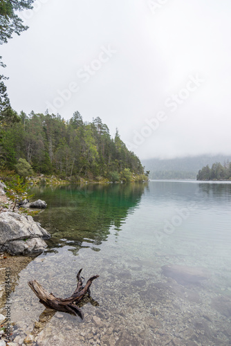 View over the Eibsee to the mountains disappearing in the fog in the background. The water is clean and clear, the shores are lined with dense forest. In the foreground is a branch in the lake.
