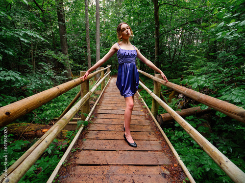 Young teenager girl in high fashion blue dress posing on a wooden bridge in a park. Prom or senior party theme. Young lady wearing beautiful outfit. Model with slim body type. Design cloths showcase