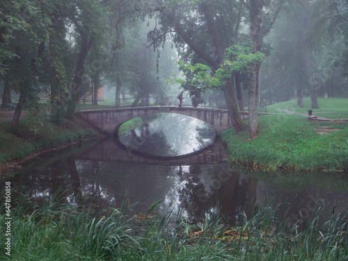 Small concrete old bridge over a small river in a town forest park in a fog. Mistry surreal calm mood. Relaxing atmosphere and melancholic nature vibe. photo