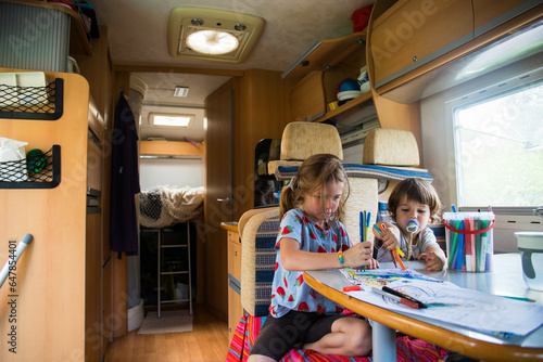 Two caucasian children, brother and sister, drawing inside a campervan during a road trip stop. Camper vacations travelling with kids background.