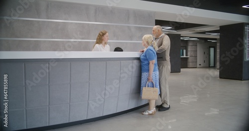 Elderly couple stand near reception desk in clinic lobby area and make appointment with doctor. Female administrator talks with patients at information counter. Medical staff work in modern hospital.