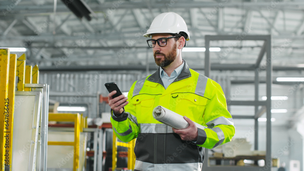 Main engineer monitors work process. Man in protective glasses and reflective clothes is holding phone. Worker with blueprint. Male is going at workshop on background of workflow and equipment. Main engineer monitors work process. Man in protective glasses and reflective clothes is holding phone. Worker with blueprint. Male is going at workshop on background of workflow and equipment.
