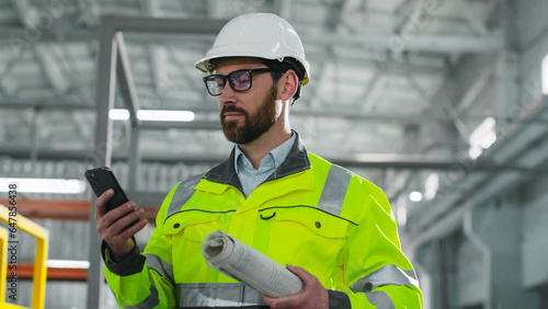 Bearded architect inspect ndustrial site. Male holds blueprint and look at phonescreen. Industrial man in reflective vest and hard hat at construction site. Worker at background of work process. photo