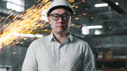 Portrait of Asian male engineer looking straight at camera and blinking. Industrial manager in uniform and safety helmet at backgrounf of welding sparks flying. Employee in googles works at workshop. © ihorvsn