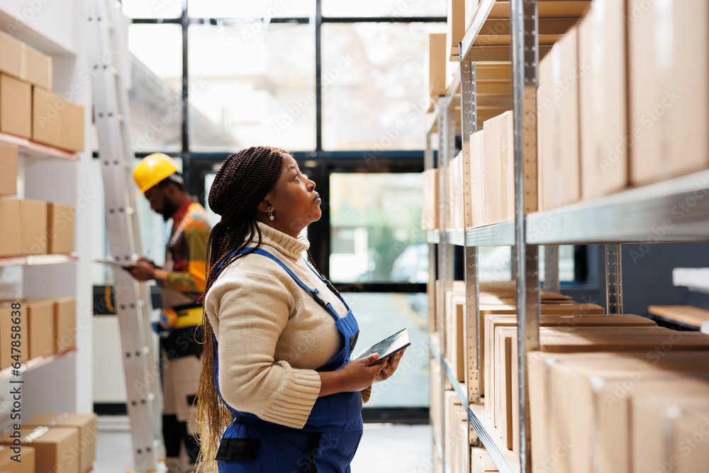 Logistics and distribution manager inspecting merchandise storage in warehouse. African american storehouse employee standing near parcels shelf, holding tablet and looking at carton Logistics and distribution manager inspecting merchandise storage in warehouse. African american storehouse employee standing near parcels shelf, holding tablet and looking at carton