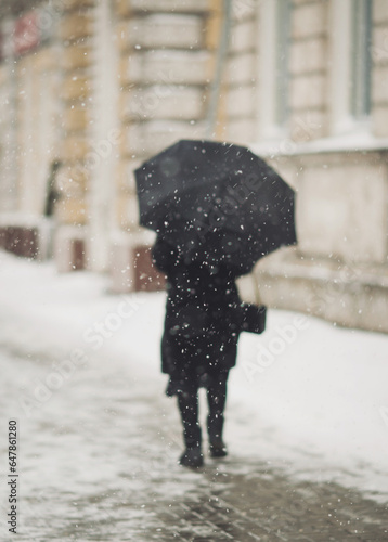 Blured figure of old woman with umbrella in warm clothes walking in the street in snowy winter day. First snow. Spending time alone in the city. Back view. figure of lonely woman going away.