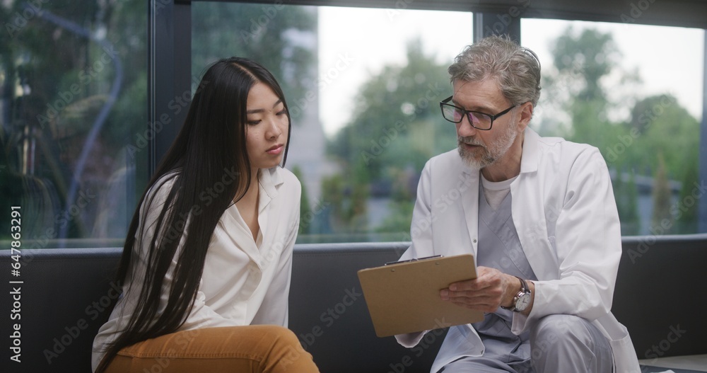 Fototapeta premium Asian woman sits on couch in clinic lobby area after appointment with doctor. Doctor talks with patient about medical examination or test results. Waiting room in medical center. Healthcare system.