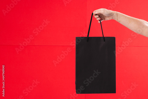 Hand of caucasian woman holding black gift bag with copy space on red background