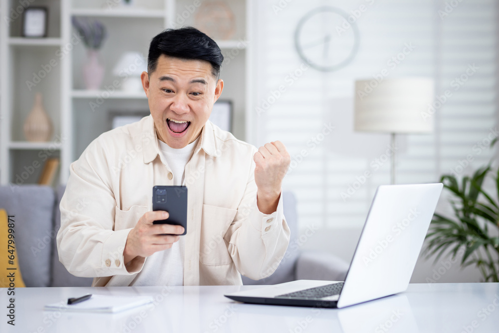 The winner is sitting at the table at home, the man is happy to receive the online notification of the victory and success on the phone, the Asian man is holding his hand up and holding smartphone. The winner is sitting at the table at home, the man is happy to receive the online notification of the victory and success on the phone, the Asian man is holding his hand up and holding smartphone.