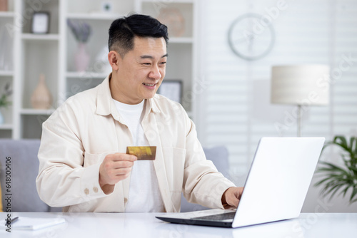 Joyful successful asian businessman working from bright home office, man using bank credit card and laptop for online shopping in online store, and booking services remotely.