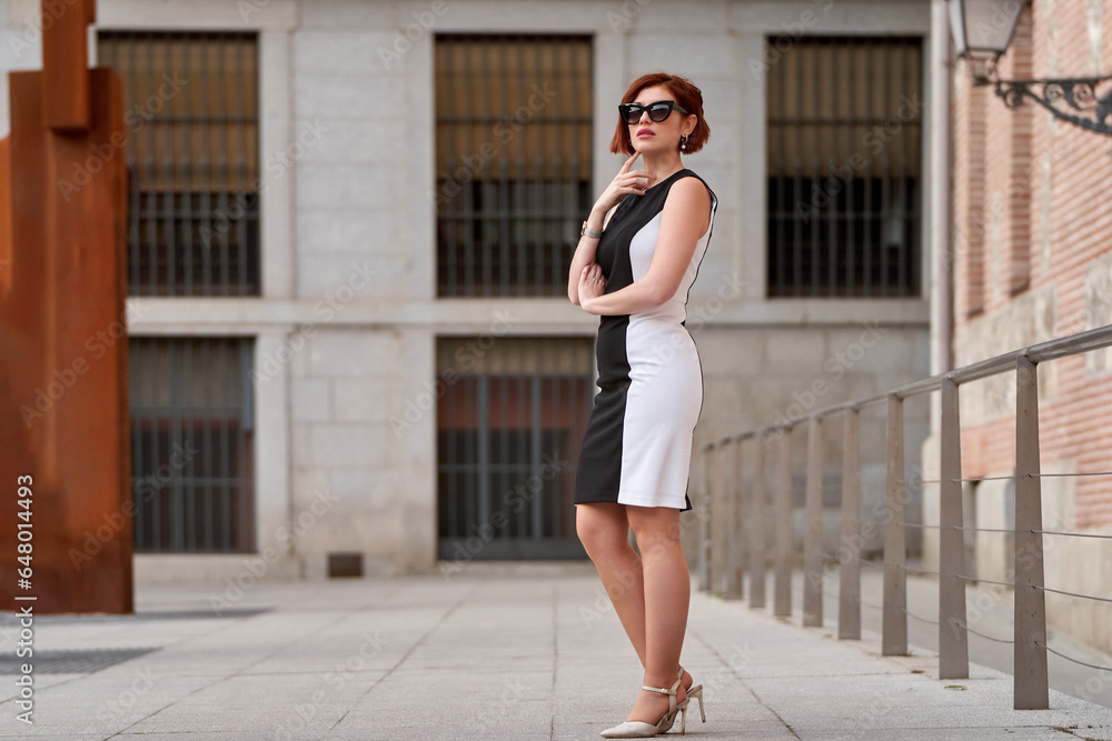 A confident and stylish redheaded woman poses for the camera, looking above her sunglasses with an alluring gaze, dressed in an elegant outfit that exudes sophistication and charm
