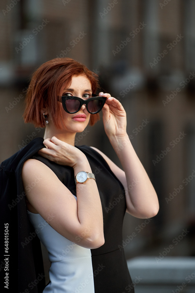 A confident and stylish redheaded woman poses for the camera, looking above her sunglasses with an alluring gaze, dressed in an elegant outfit that exudes sophistication and charm A confident and stylish redheaded woman poses for the camera, looking above her sunglasses with an alluring gaze, dressed in an elegant outfit that exudes sophistication and charm