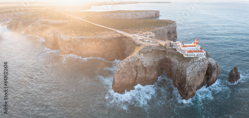 Atop the rugged cliffs of Cabo de São Vicente, Farol do Cabo de São Vicente lighthouse, panoramic view of the Atlantic Ocean, making it a visit place on vacations in the Algarve region. Aerial view.