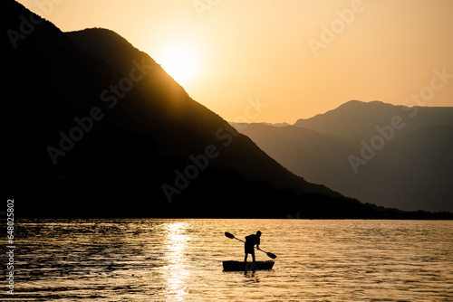 At sunset, amidst the mountains, a fisherman rows a small boat in the bay. Silhouettes in the golden hour.