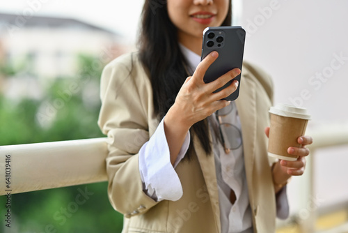 Smiling businesswoman texting messages, chatting in social on smartphone while standing on balcony