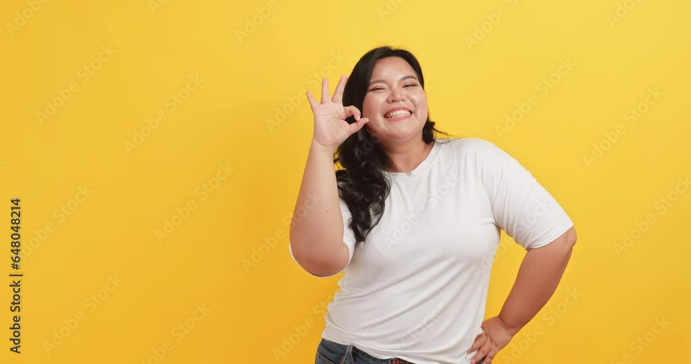 A chubby Asian woman wearing a white t-shirt makes a gesture of agreement and satisfaction with something on a yellow background. A chubby Asian woman wearing a white t-shirt makes a gesture of agreement and satisfaction with something on a yellow background.