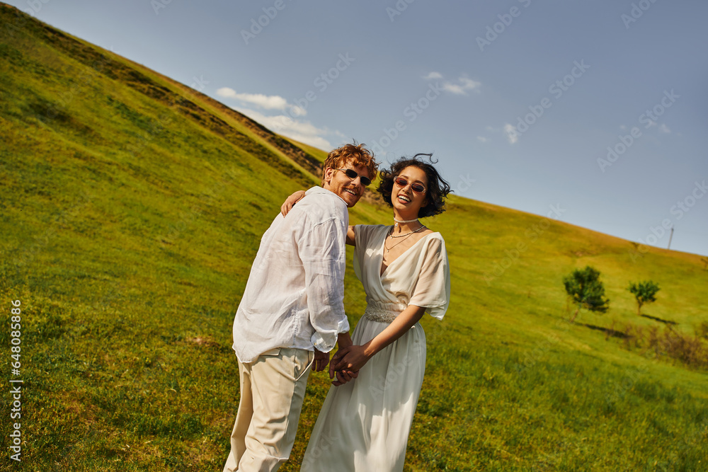 rural wedding in countryside, multiethnic newlyweds in wedding gown looking at camera in green field rural wedding in countryside, multiethnic newlyweds in wedding gown looking at camera in green field