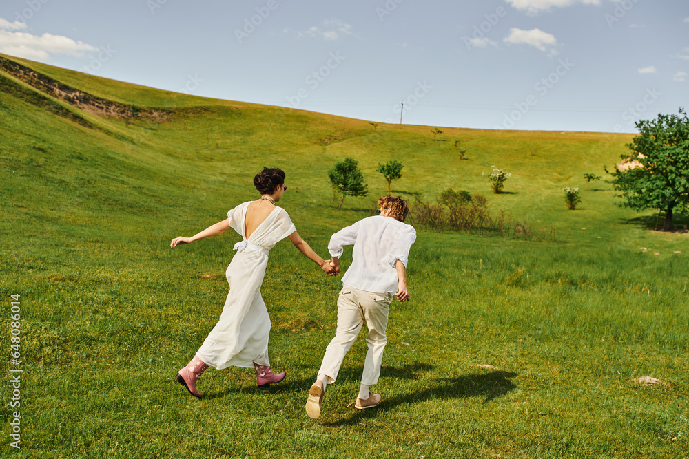 back view of couple of newlyweds running in green field, rustic wedding, boho style white attire back view of couple of newlyweds running in green field, rustic wedding, boho style white attire