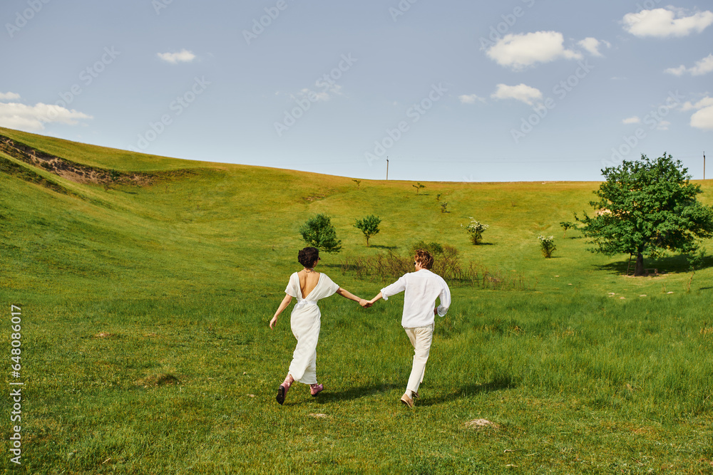 young couple of newlyweds running in green field, bride and groom in wedding gown holding hands young couple of newlyweds running in green field, bride and groom in wedding gown holding hands